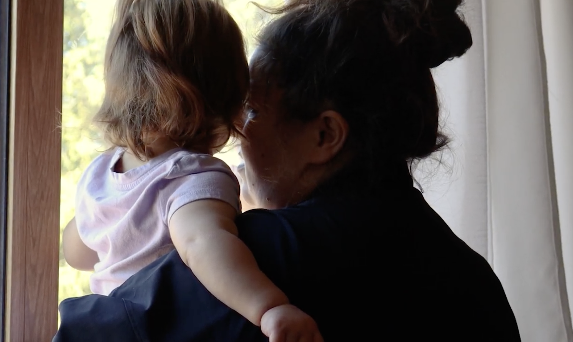 This picture has an adult woman holding a toddler looking outside a window
