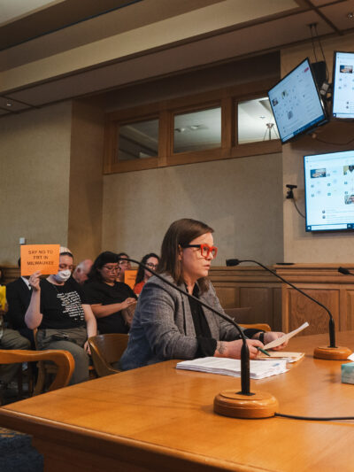 Policy Director Amanda Merkwae testifying in front of the Milwaukee Equal Rights Commission and protesters holding signs reading "No FRT in MKE"