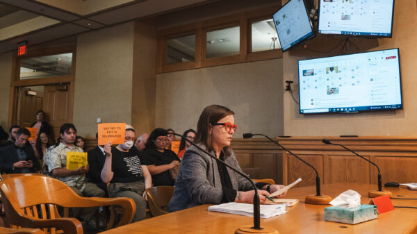 Policy Director Amanda Merkwae testifying in front of the Milwaukee Equal Rights Commission and protesters holding signs reading "No FRT in MKE"
