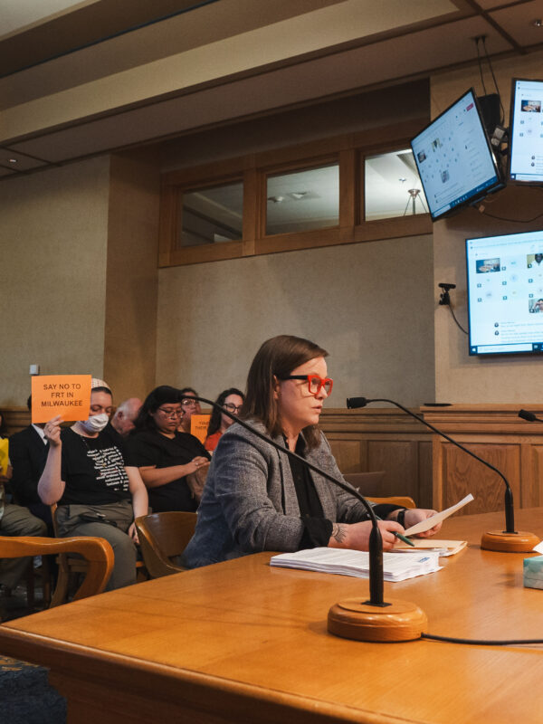 Policy Director Amanda Merkwae testifying in front of the Milwaukee Equal Rights Commission and protesters holding signs reading "No FRT in MKE"